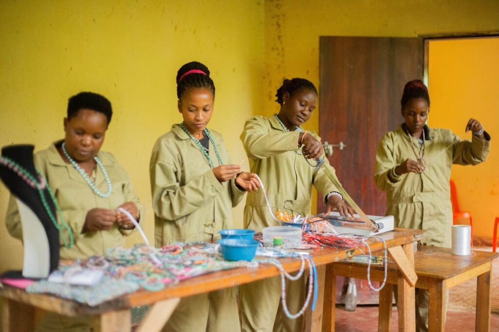 Women in uniforms creating beaded jewelry in an indoor craft workshop.